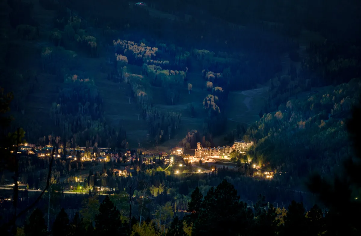 A twilight view featuring the twinkling lights of a cozy mountain town, surrounded by dense forest and set against a dramatic mountain backdrop. The nighttime atmosphere evokes peaceful evenings and the allure of stargazing or night walks in a pristine alpine setting.