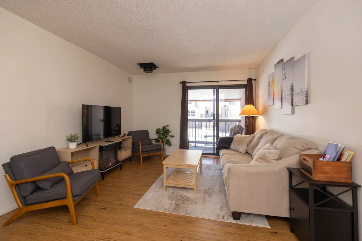 The image showcases a cozy, inviting living area with a plush beige sofa adorned with cushions, two stylish gray armchairs with wooden accents, a flat-screen TV on a wooden stand, and a sliding glass door that opens onto a balcony with potential mountain views.