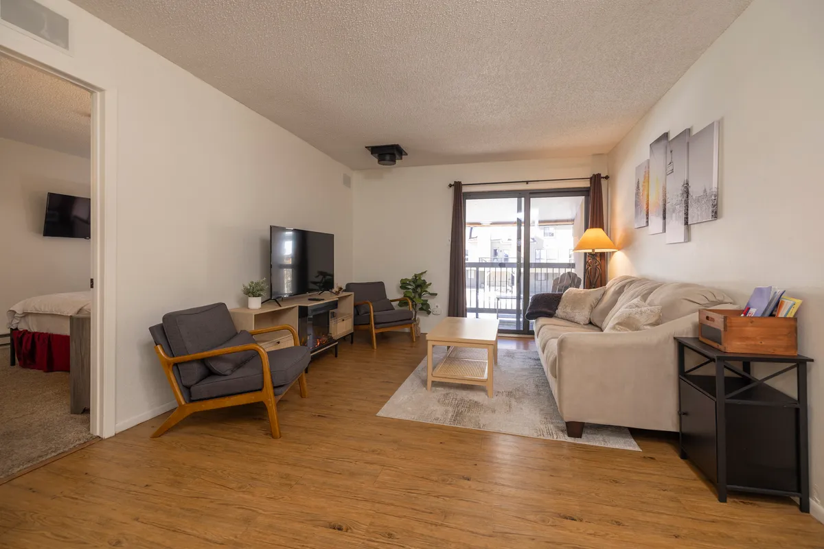 A cozy living room scene with a beige sofa, two modern armchairs, and a flat-screen TV on a media console. A sliding glass door leads to a balcony, suggesting openness and flexibility. This space can double as an additional sleeping area with the sleeper sofa for extra guests.