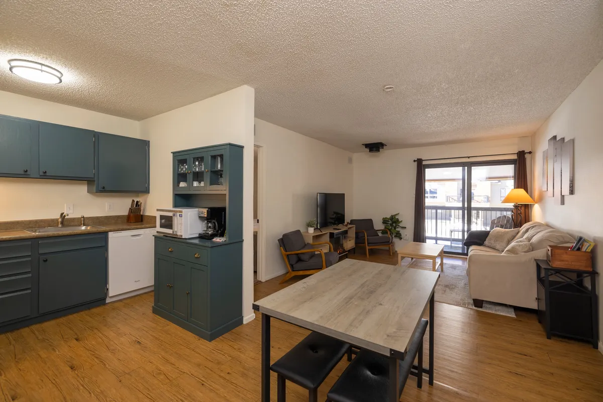 A wide-angle interior shot capturing the open-concept living area, which includes the teal kitchen cabinetry, a dining table with benches, and a comfortable living room seating arrangement. The space is well-lit by natural light coming through the balcony door, highlighting the seamless flow between the kitchen, dining, and lounging areas.