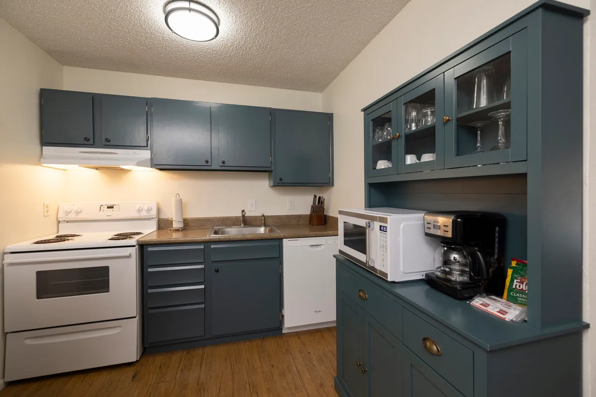 A cozy condo kitchen featuring blue cabinets with brass knobs, a white electric stove and oven, a microwave and coffee maker on a blue hutch, and glassware displayed in the upper cabinet. The space feels inviting and practical, perfect for preparing meals or hot drinks after a day on the slopes.