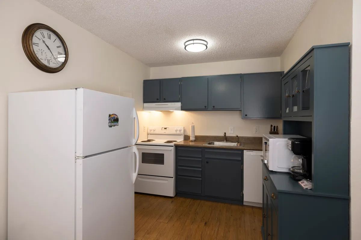 A focused view of the kitchen area highlighting blue cabinets, a white refrigerator, stove, oven, and wall clock. The setup is practical yet inviting, designed for meal prep, casual family interactions, and sharing stories around the kitchen.