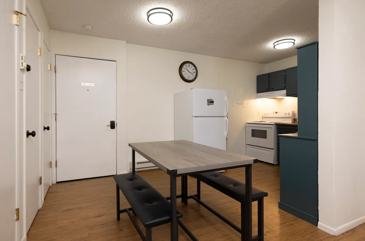 A cozy, practical dining area with a wooden table and benches, a white refrigerator, stove, and wall clock, suggesting a welcoming spot for shared meals and planning sessions after a day outdoors.