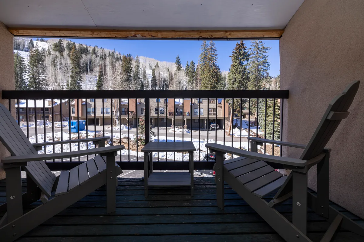 Two Adirondack chairs sit on a balcony overlooking a snow-covered slope and neighboring townhouses. The inviting setup suggests a peaceful spot to relax after a day of adventure, enjoying the crisp mountain air and scenic views.