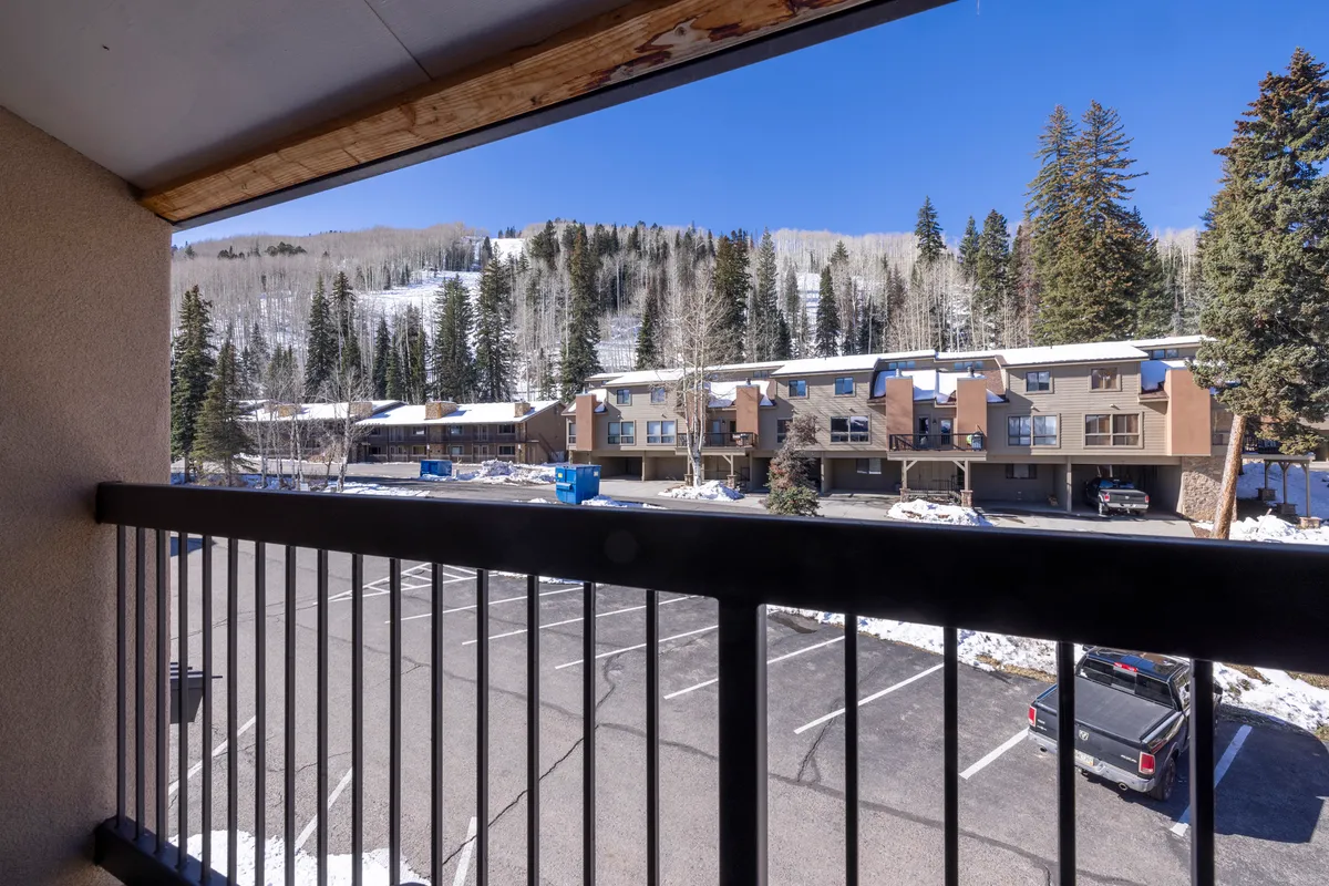 A peaceful winter scene from a balcony, looking out over a snow-covered parking lot and townhome-style buildings, with tree-lined mountain slopes forming a striking backdrop. The balcony railing frames the inviting view, evoking a sense of retreat and connection with the mountains.