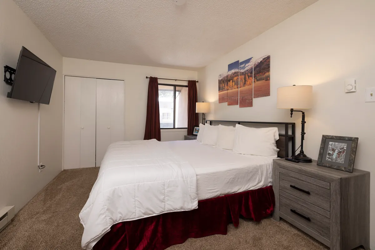 A calm, inviting master bedroom with a king bed dressed in white bedding and a red bed skirt. Above the bed, a triptych of alpine wall art adds a mountain retreat feel. The room also features matching bedside tables with lamps and a wall-mounted flat screen TV, emphasizing relaxation and comfort.