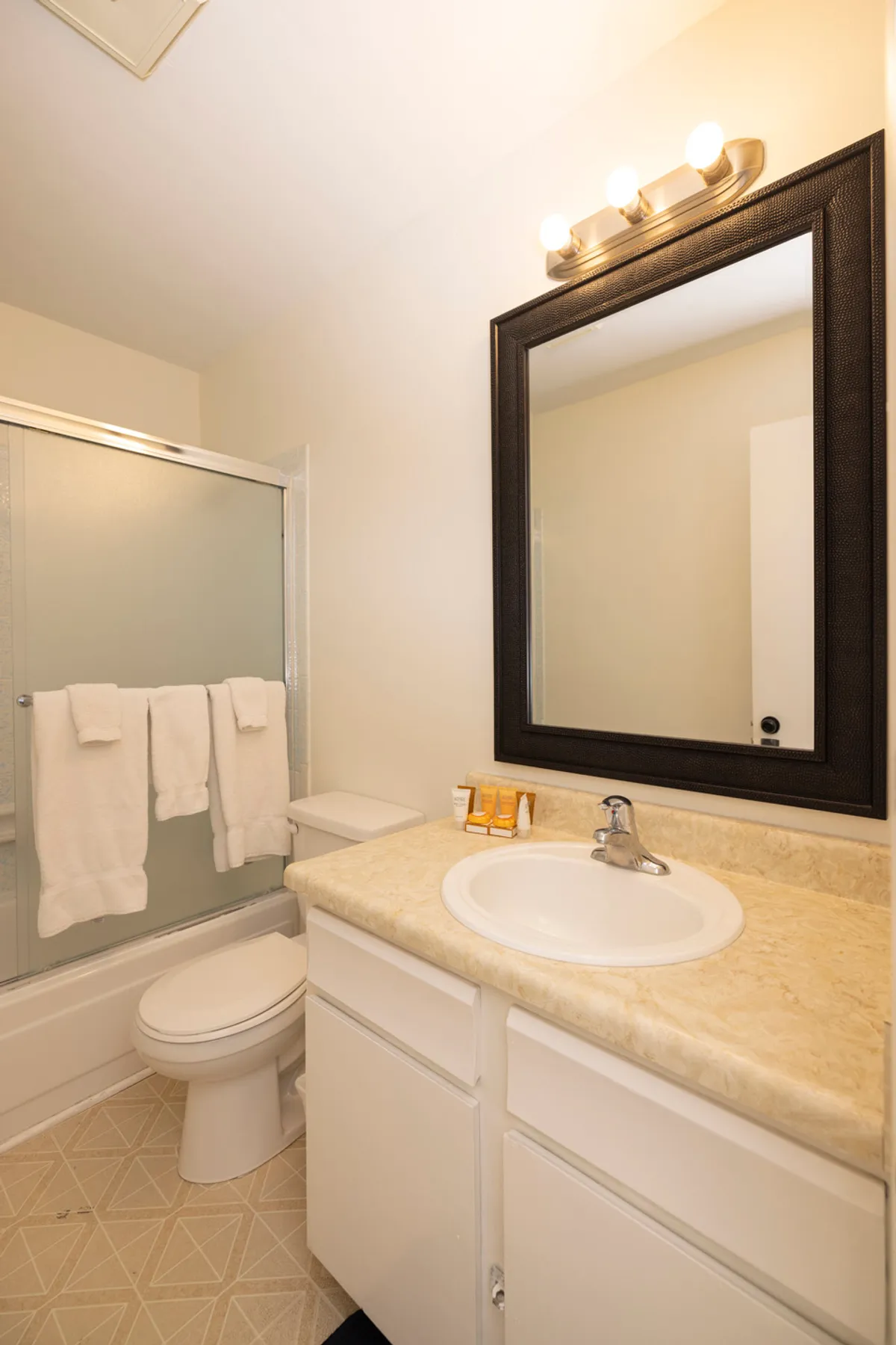 A practical bathroom scene with a sink set in a marble countertop, a large dark-framed mirror, a toilet, and a shower area with a frosted glass sliding door and towel rack. The space feels cozy and functional, perfect for daily routines.