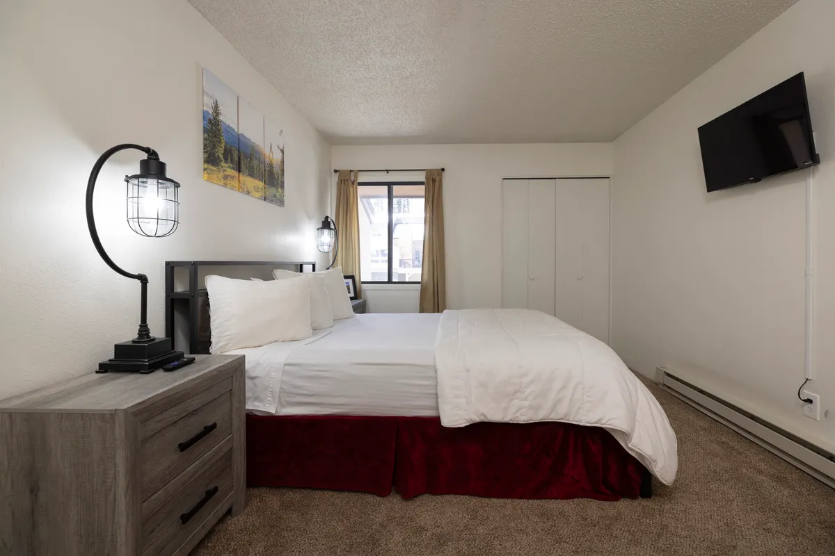 Another angle of the master bedroom, featuring a king bed with white linens and red bed skirt. The room is equipped with modern bedside lamps, a wall-mounted TV, and a window with beige curtains, providing a glimpse of the exterior building and natural light.
