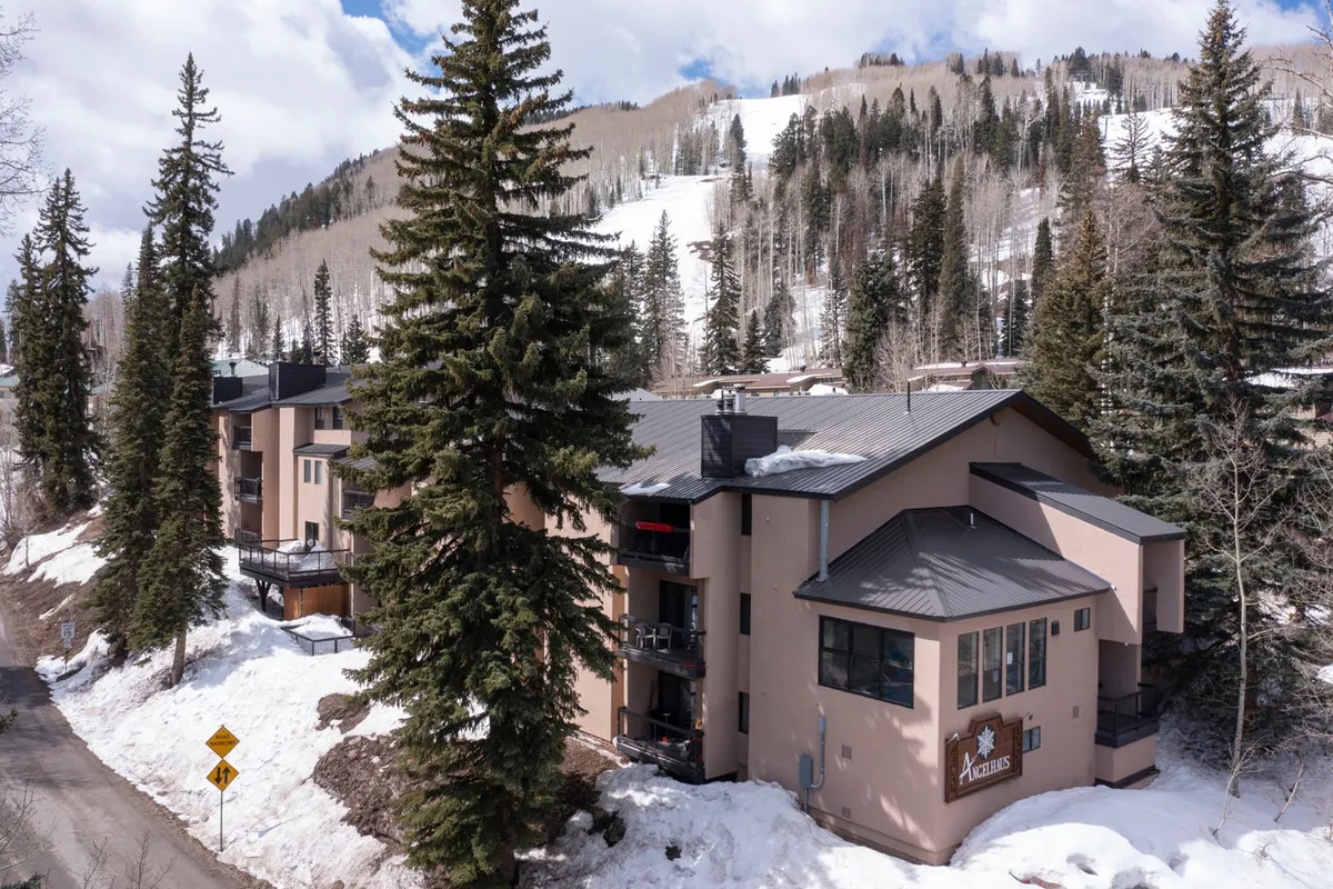 This image shows the Angel Haus building surrounded by tall evergreen trees, with snow-covered ski slopes visible in the background. The balconies face toward the mountains, suggesting a peaceful retreat with easy access to the outdoors. The overall scene is tranquil and inviting, with a winter getaway ambiance.