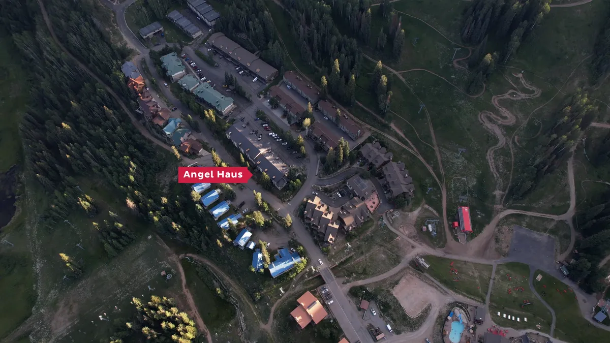This exterior shot showcases the Angel Haus buildings with their distinctive blue roofs, set amid a surrounding pine forest. Visible nearby are ski slopes and winding trails, as well as the community swimming pool area, encapsulating the property's proximity to both mountain adventure and leisure facilities.