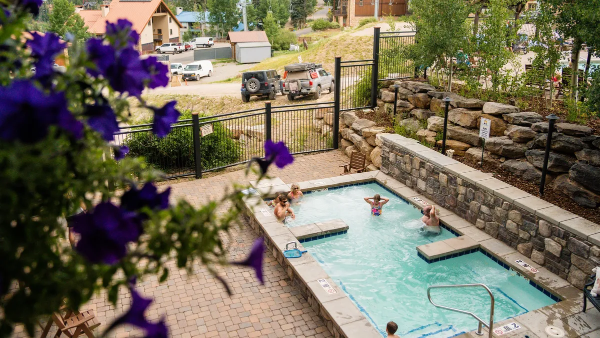 An inviting view of the heated community swimming pool surrounded by tasteful landscaping, a stone wall, and blooming purple flowers. Families and friends are pictured enjoying the water, highlighting the cheerful and relaxing atmosphere of the pool area.