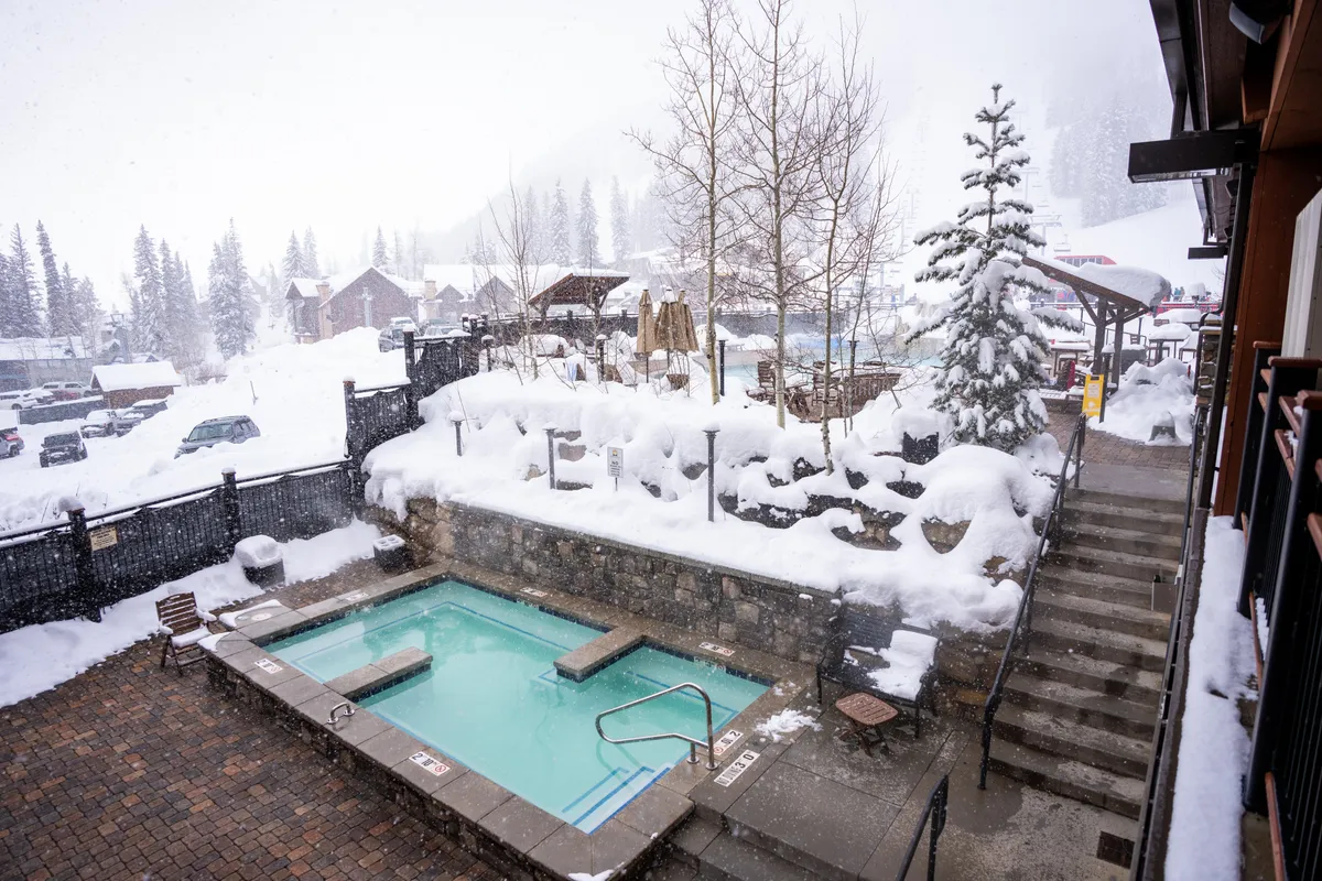 A heated outdoor swimming pool with clear aqua water is set against a backdrop of snow-laden pine trees and alpine buildings. A snow-covered stone retaining wall and a staircase leading to an upper patio area provide depth and context. The scene highlights the unique experience of swimming in a heated pool while surrounded by a winter landscape.