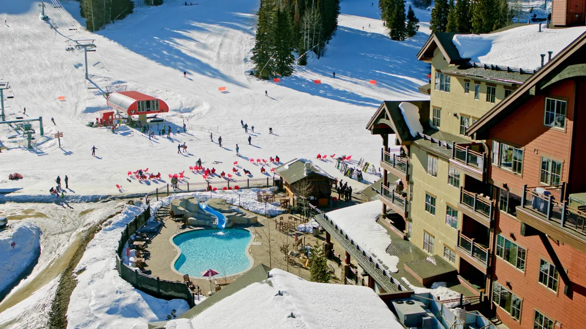 A vibrant exterior scene featuring snow-covered ski slopes dotted with skiers and snowboarders, a red chairlift station, and a heated outdoor swimming pool with a water slide. The multi-story alpine lodge with balconies overlooks all the action, blending active winter fun with relaxation.