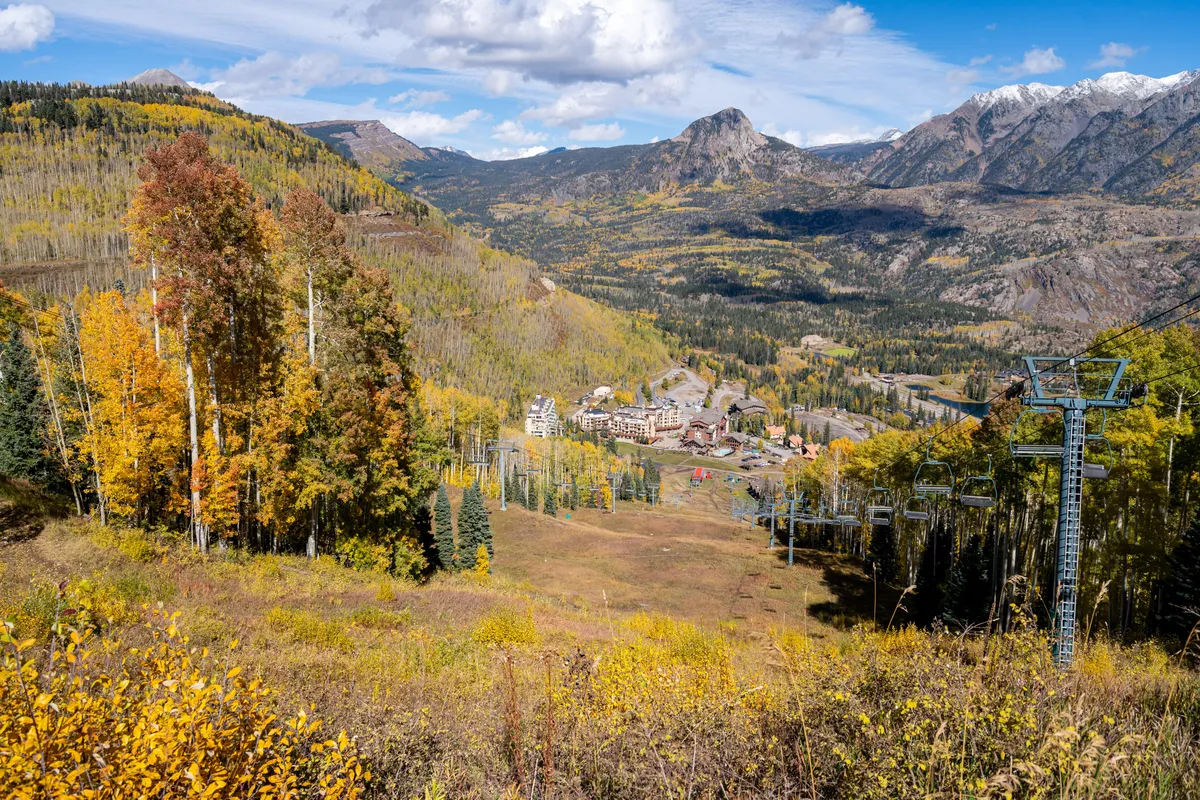 The scene showcases a ski lift rising toward light-dusted mountain peaks, with a cluster of buildings nestled at the valley base. Colorful autumn trees add seasonal vibrancy, highlighting both winter and summer exploration opportunities.