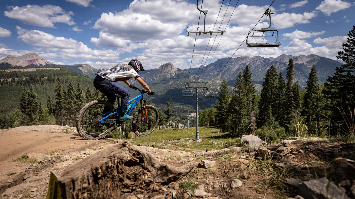 A mountain biker is captured mid-jump on a dirt trail, with ski lift chairs in the background and a dramatic alpine mountain range rising behind. Dense pine forests frame the scene, highlighting the property's immediate access to both summer and winter outdoor adventures.