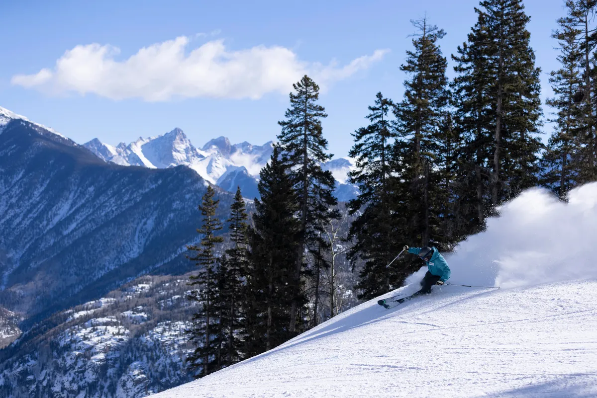 A skier carves down a pristine, snow-covered slope framed by tall pine trees and distant, rugged mountain peaks. The scene captures the excitement of alpine adventure at the doorstep, with easy access to skiing and snowboarding activities.